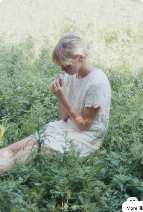 a young woman with blonde hair pulled back sits gracefully in a field of lush green grass. she is wearing a delicate white lace dress and is gently holding a small flower in her hands. her head is slightly bowed, as if she is captivated by its simple beauty. the overall atmosphere is one of tranquility and peace, bathed in the warm glow of natural sunlight.