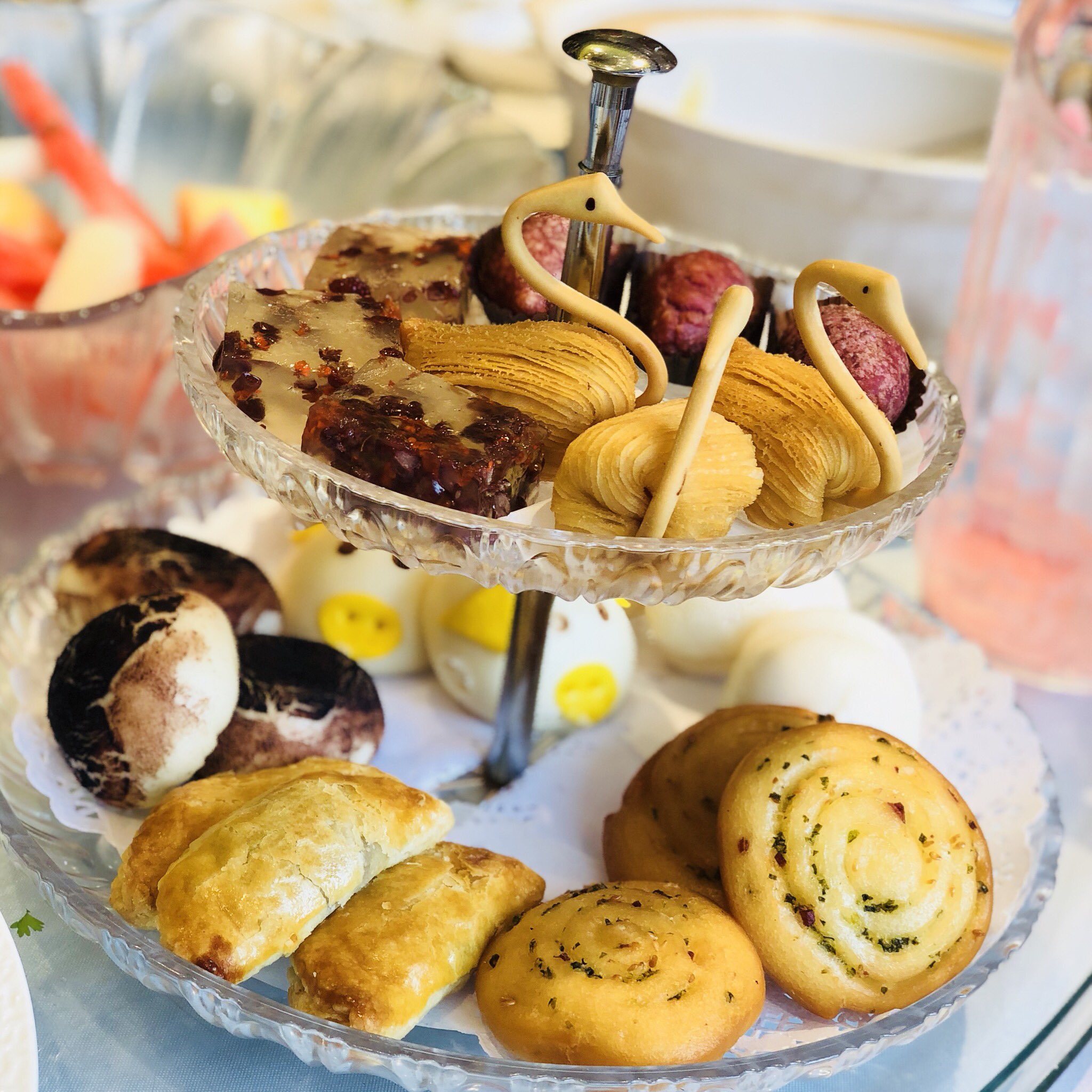 a beautiful tiered serving tray piled high with a variety of chinese pastries and dim sum. the top tier features intricately shaped swan pastries and translucent jelly desserts. the bottom tier holds mushroomshaped pastries, golden buns, and savory rolls. the table setting is simple, allowing the colorful and artistic food to take center stage.