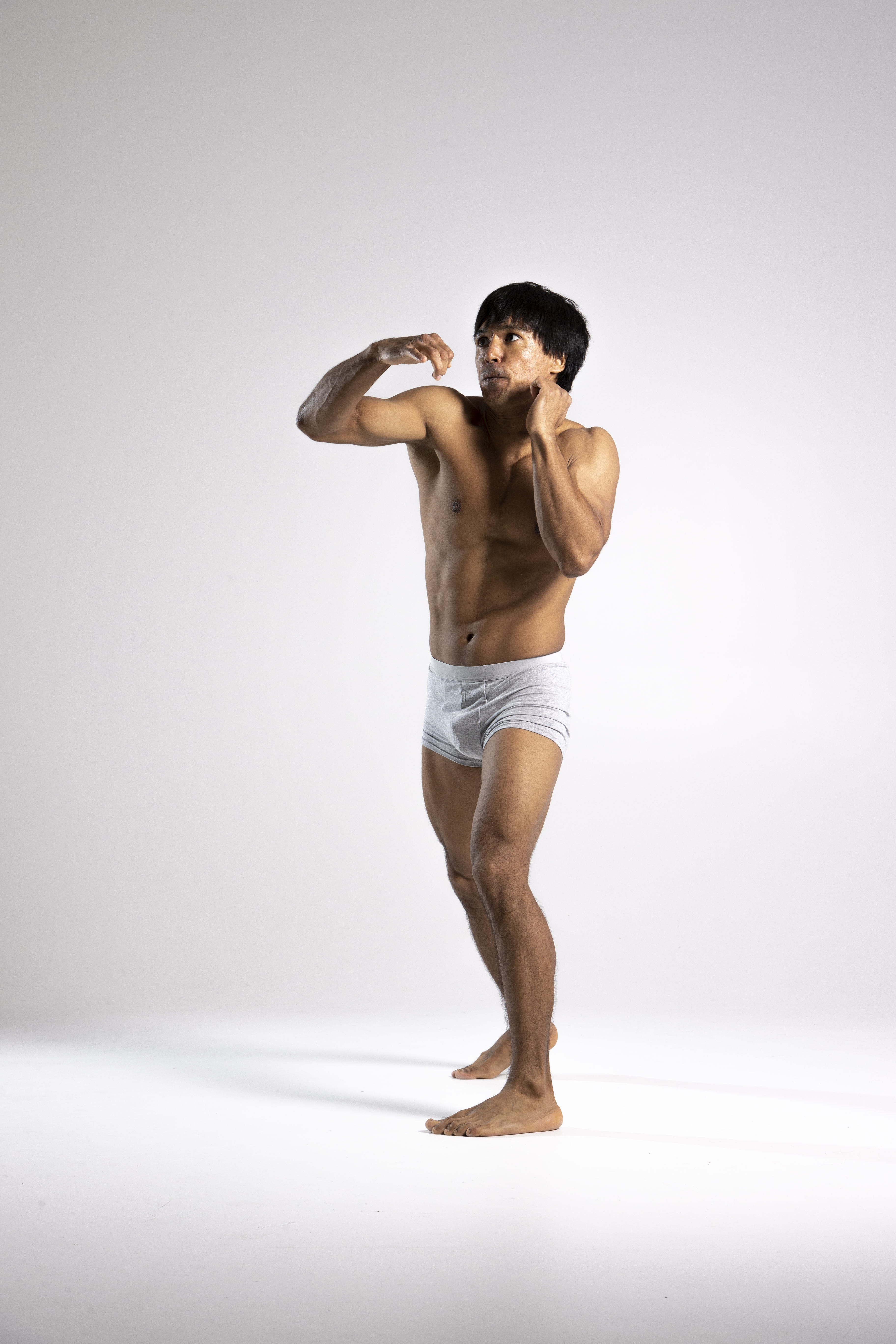 a full shot of a shirtless, muscular man wearing briefs, standing barefoot on a white background, looking away with a defensive pose.