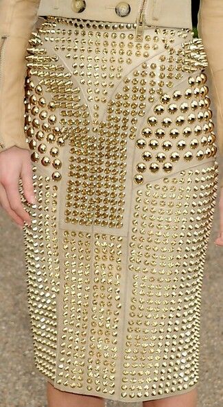 a closeup image of a beige skirt densely covered in gold studs of varying sizes and shapes. the studs are arranged in patterns, and the fabric of the skirt appears to be a structured material. the image focuses on the texture and design of the garment.