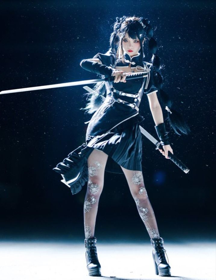 a young woman in a black dress, skirt, and tights is holding two swords. she is posing in a dramatic fashion, with her hair flowing behind her. the photo is taken in a dark studio with a few lights shining on her.