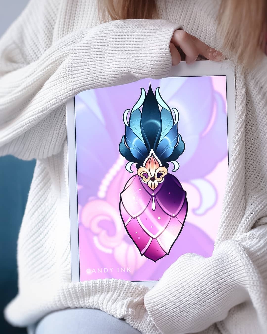 a woman in a white sweater holds a tablet displaying a digital drawing of a heartshaped tattoo design. the heart features a stylized, scalelike pattern in shades of pink and purple, topped with a blue and teal floral ornament and a gold fleurdelis. the background of the drawing is a soft pink and purple gradient. the image is brightly lit and taken from a straighton perspective.