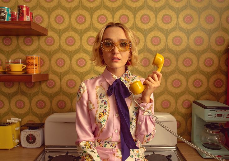 a woman in a pink and floral dress and yellow glasses stands in a retro kitchen talking on a rotary phone. the kitchen has floral wallpaper and a yellow and green color scheme. she appears to be in her 20s or 30s.