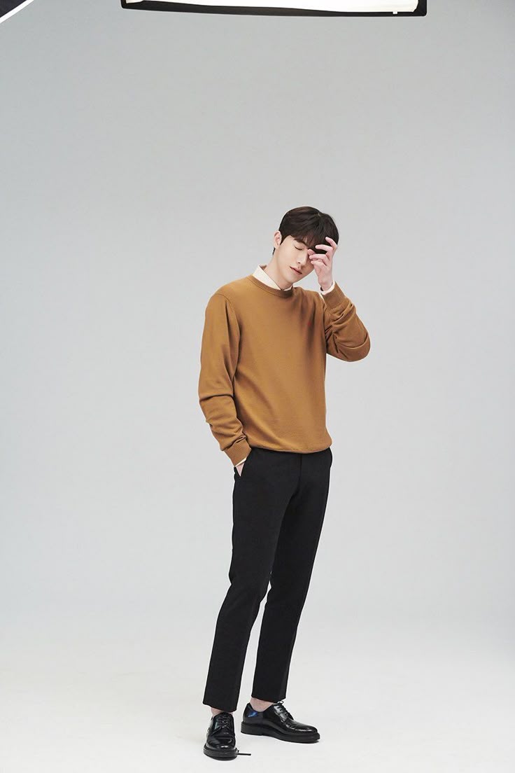 a full shot shows a man in a brown sweater, black pants, and black dress shoes in a studio. he is standing with one hand in his pocket and the other on his face.