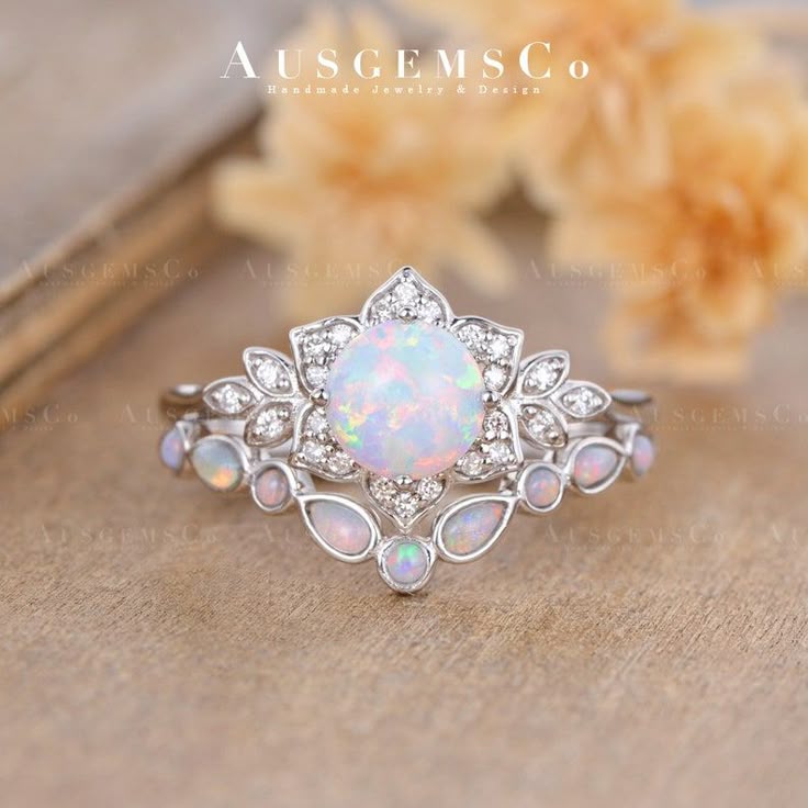a closeup shot showcases a white gold engagement ring set featuring a large round opal at its center, surrounded by small diamonds. the design includes a decorative florallike setting, and the ring is accompanied by a matching opal wedding band with small opal accents. the image is likely an advertisement for handmade jewelry by ausgemsco, emphasizing its vintageinspired and luxurious design. the rings are displayed on a textured surface with blurred flowers in the background, suggesting a bridal or special occasion theme.