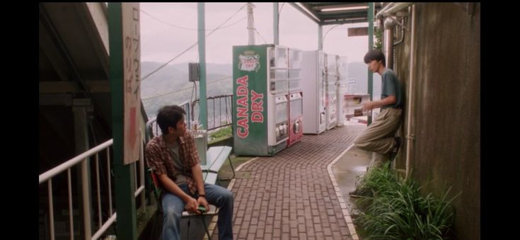 two young asian men, one sitting and one standing, are seen in the foreground of this outdoor shot.  they appear to be friends, enjoying a quiet moment.  behind them are several vending machines, including one dispensing canada dry ginger ale. the setting is outdoors, on a brick walkway, with a scenic view of mountains in the background. the overall atmosphere is calm and relaxed, suggesting a summer day. the clothing of the young men is casual.