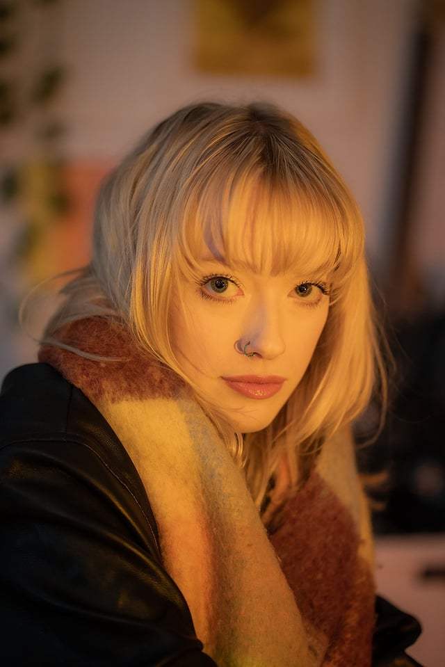 closeup portrait of a young woman with blonde hair and bangs. she is wearing a black leather jacket and a multicolored scarf. the lighting is warm and the background is blurred. the woman is looking directly at the camera.