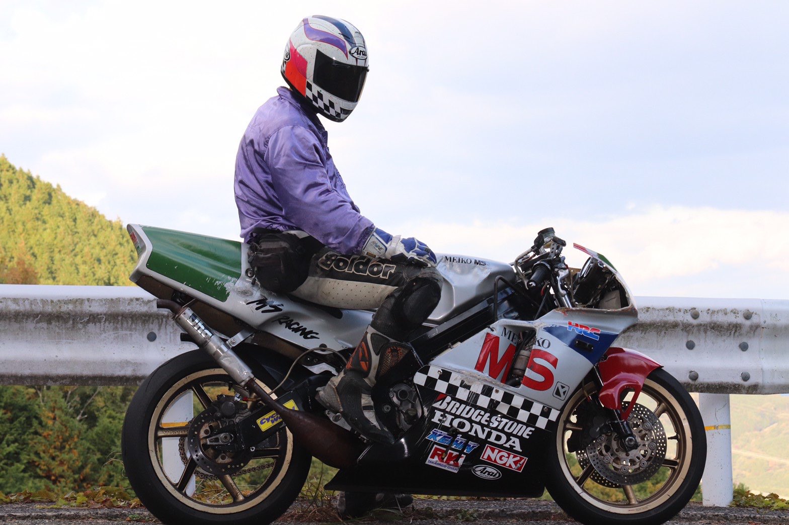 a person wearing a motorcycle helmet and riding a motorcycle on a road. the motorcycle is a silver and red honda. the rider is wearing a purple jacket and white pants.  the motorcycle is parked near a guardrail and a mountain forest.  the motorcycle has a bridge stone tire and a honda logo. the rider has a helmet and riding gear.
