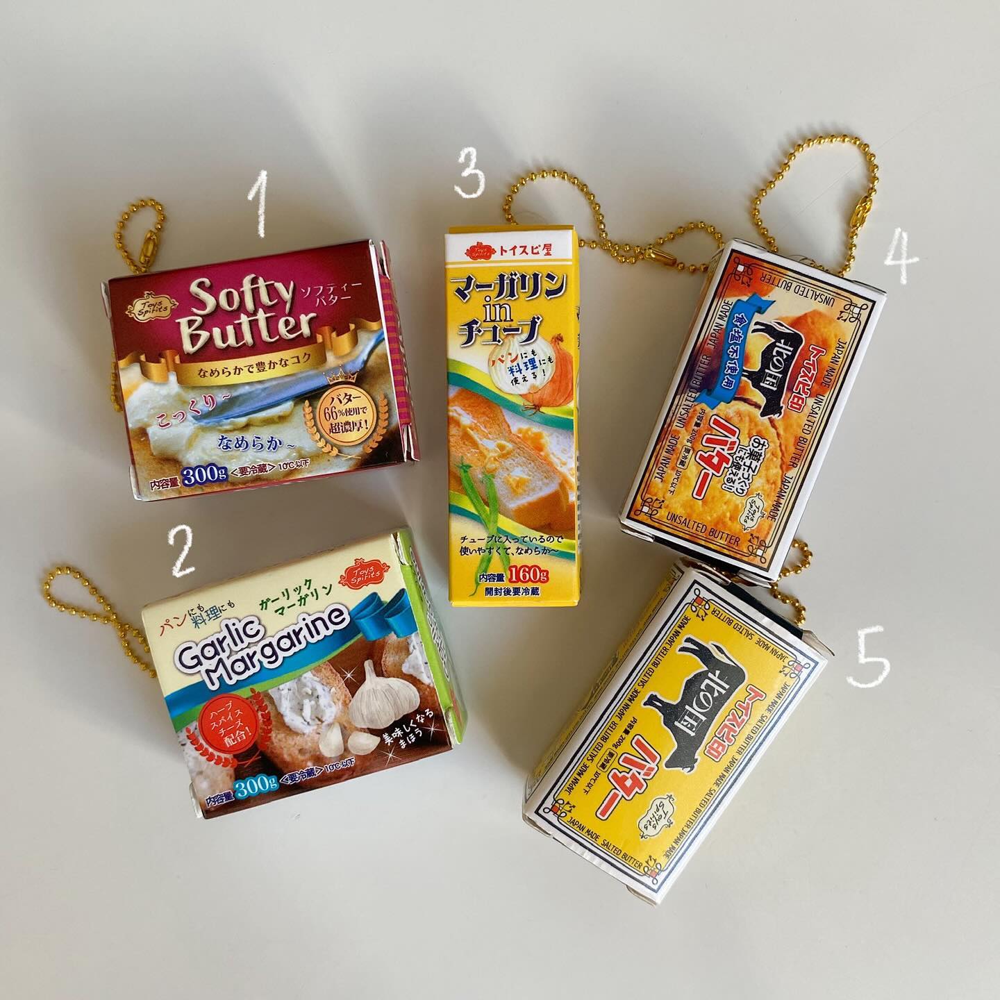 the image shows five miniature butter and margarine boxes, labeled with numbers 1 through 5. they seem to be keychains, as they are each attached to a small golden chain. the products appear to be japanese, with writing in japanese characters. there are boxes of "softy butter", "garlic margarine", "margarine in tube" and two boxes of "unsalted butter". the background is a plain white surface.