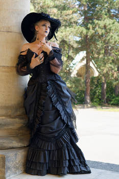 a woman in a black victorianera gothic dress stands against a stone column in a garden setting. the dress is long, with multiple layers of ruffles and a corset bodice.  she wears a widebrimmed black hat. the overall scene evokes a dark, mysterious and elegant mood.  the background includes mature trees and greenery.