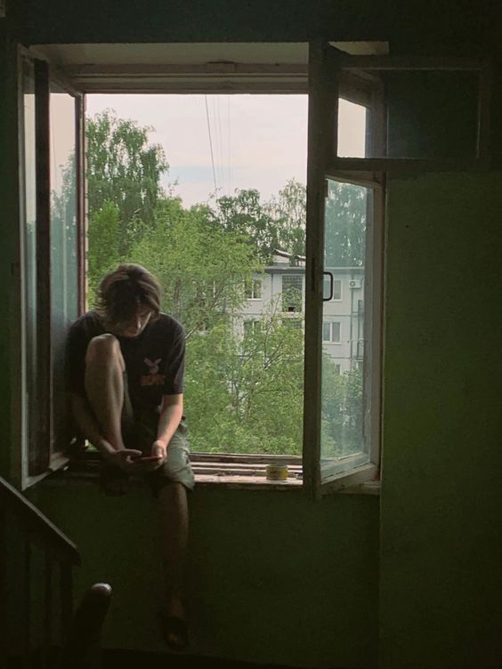 a young man sits on the windowsill of an open window, his back to the room. he is bathed in soft, natural light coming through the window and is looking down at his phone. outside the window, we can see green trees and a tall residential building. the overall mood is contemplative and serene.