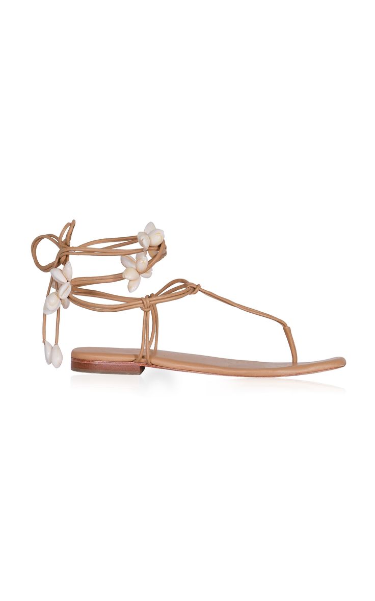 the image shows a pair of women's beige leather flat sandals.  the sandals have a minimalist design with thin straps that tie around the ankle.  the ankle straps are adorned with small, white shell embellishments. the sole of the sandal is flat and the overall style is casual and summery.