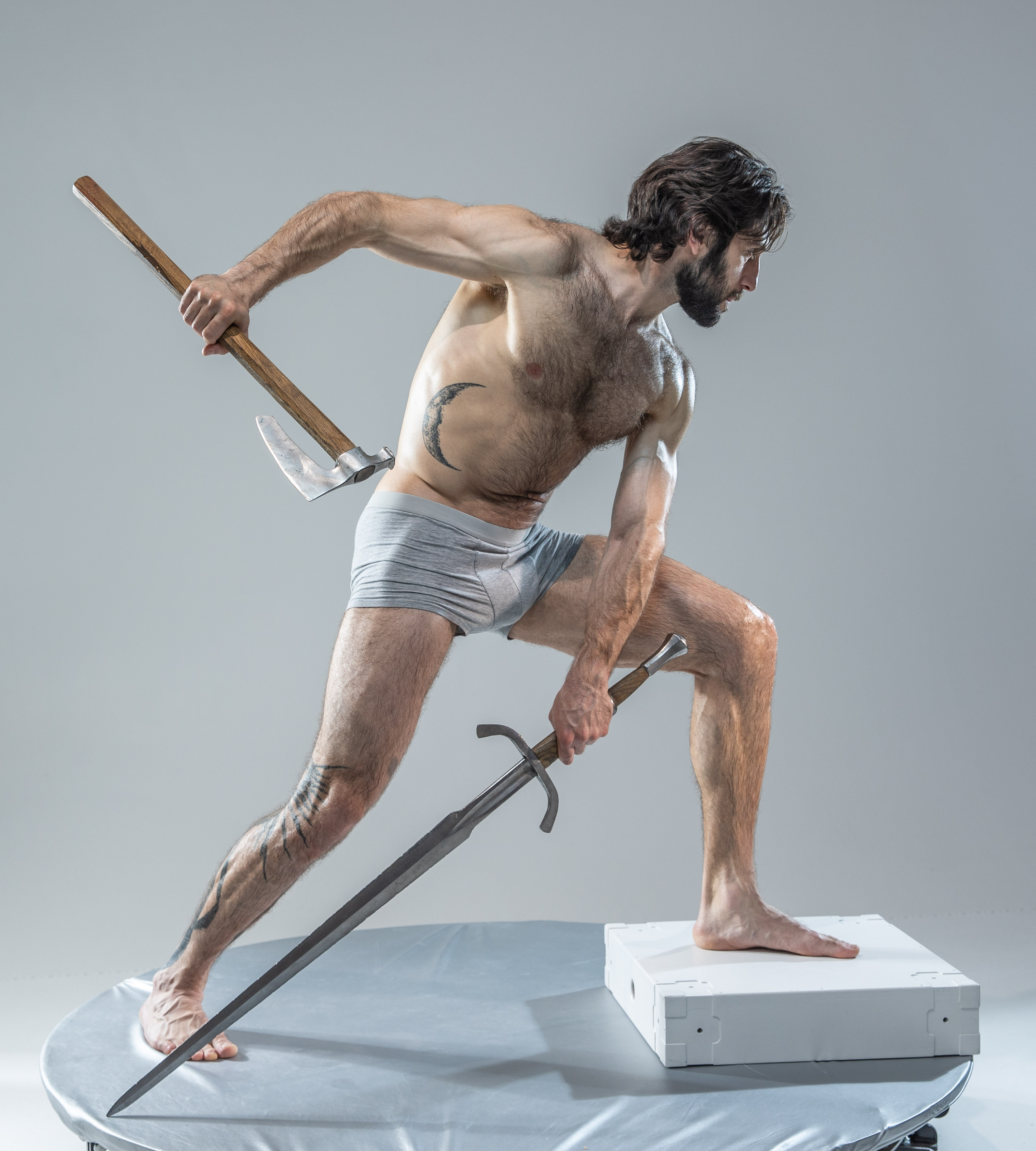 against a minimalist grey backdrop, a muscular man assumes a dynamic warrior pose. he holds a battle axe aloft in his right hand, the weapon angled downwards across his back. his left hand grips the hilt of a medievalstyle sword, its blade extending forward. dressed only in grey underwear, his toned physique is highlighted by the soft studio lighting.  the overall composition emphasizes power and athleticism,  evoking a sense of action and drama.
