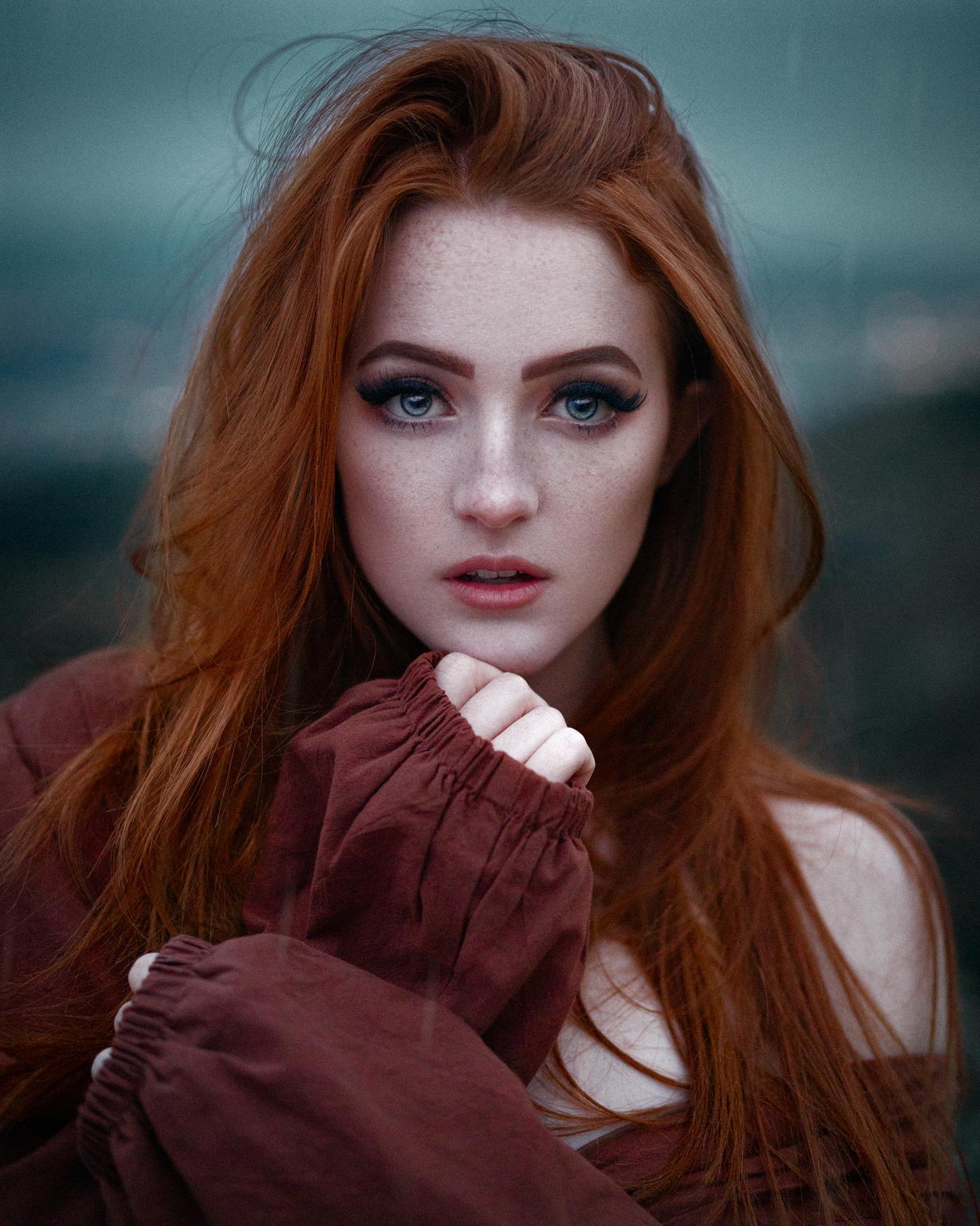 a closeup portrait of a young woman with long, flowing red hair and striking blue eyes. she has a smattering of freckles across her face, which adds to her natural beauty. she's wearing a rustcolored garment and posing with her hand gently touching her chin. the background is a blurred, moody landscape, which helps to create a sense of atmosphere and depth.