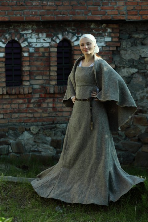 a woman is standing in front of a stone and brick building wearing a medieval style gown and headscarf. she has a belt around her waist. she is standing in a grassy area.