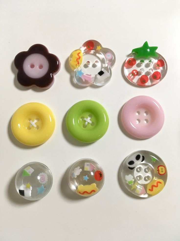 a topdown shot features nine craft buttons arranged in a 3x3 grid on a white surface. the buttons vary in shape, color, and design. the first row includes a brown flowershaped button, a clear flowershaped button with colorful cartoon details, and a clear strawberryshaped button with red and green accents. the second row consists of solidcolored circular buttons in yellow, green, and pink. the third row showcases clear circular buttons with embedded cartoon designs.