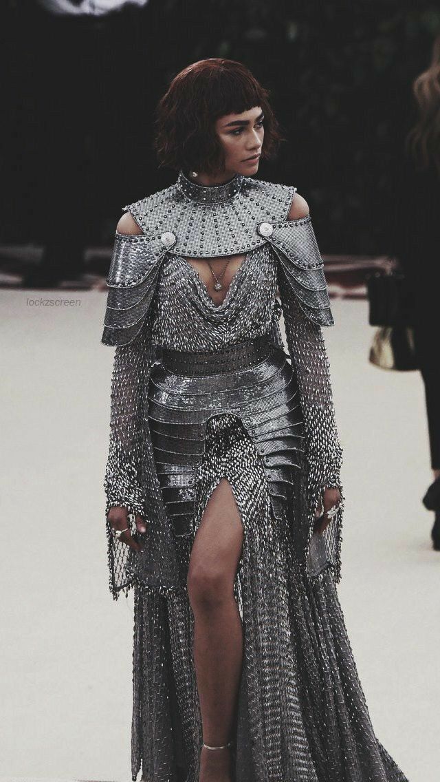 zendaya at the 2018 met gala. she is wearing a stunning silver chainmail gown that resembles armor, with a high slit.  her hair is styled in a short, blunt bob. she is posing for photos on the red carpet. the overall image has a dark, moody aesthetic.