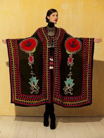 a woman is standing in front of a textured yellow wall wearing a long, black kaftan with colorful embroidery. the kaftan has a wide, rectangular shape with short sleeves, and features designs of red circles with black birdlike shapes inside, as well as turquoise leaflike patterns. the edges of the kaftan are bordered with red, yellow, and white dots. the woman has dark hair pulled back and is wearing a black turtleneck, a silver necklace with tassels, and black boots. her arms are extended to the sides, showcasing the width of the kaftan. the floor is a light beige color with shadows.