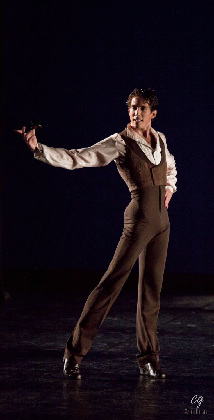 a man in a white shirt, brown pants, and vest is dancing on a stage. he has one hand up in the air and the other on his hip. he is looking to his left, as if he's focused on the audience or his partner. he has his legs bent at the knee and is in a pose that suggests movement. the lighting is dark but highlights him.
