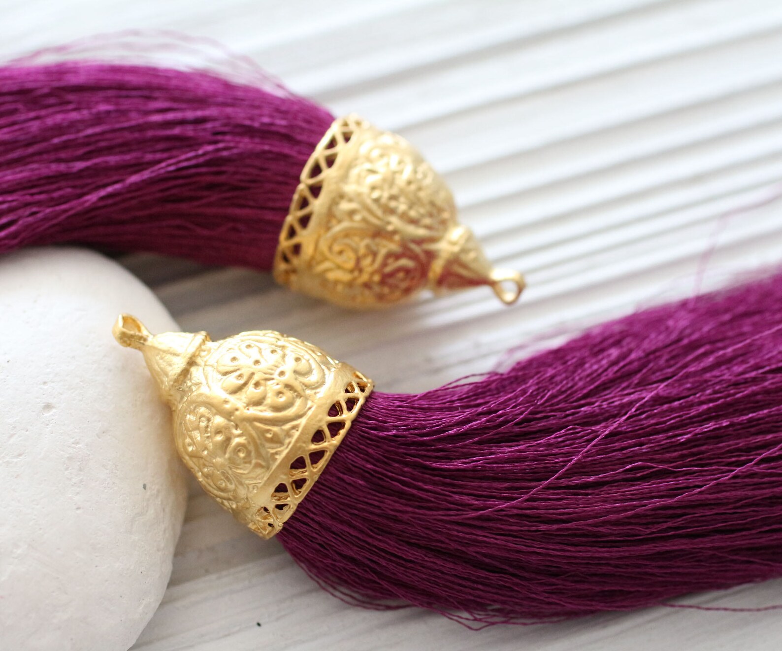 two purple silk tassels with goldplated caps are displayed on a white wooden surface. the tassels are soft and flowy, and the gold caps are intricately engraved. a white stone is partially visible in the background, adding a touch of natural elegance to the composition.