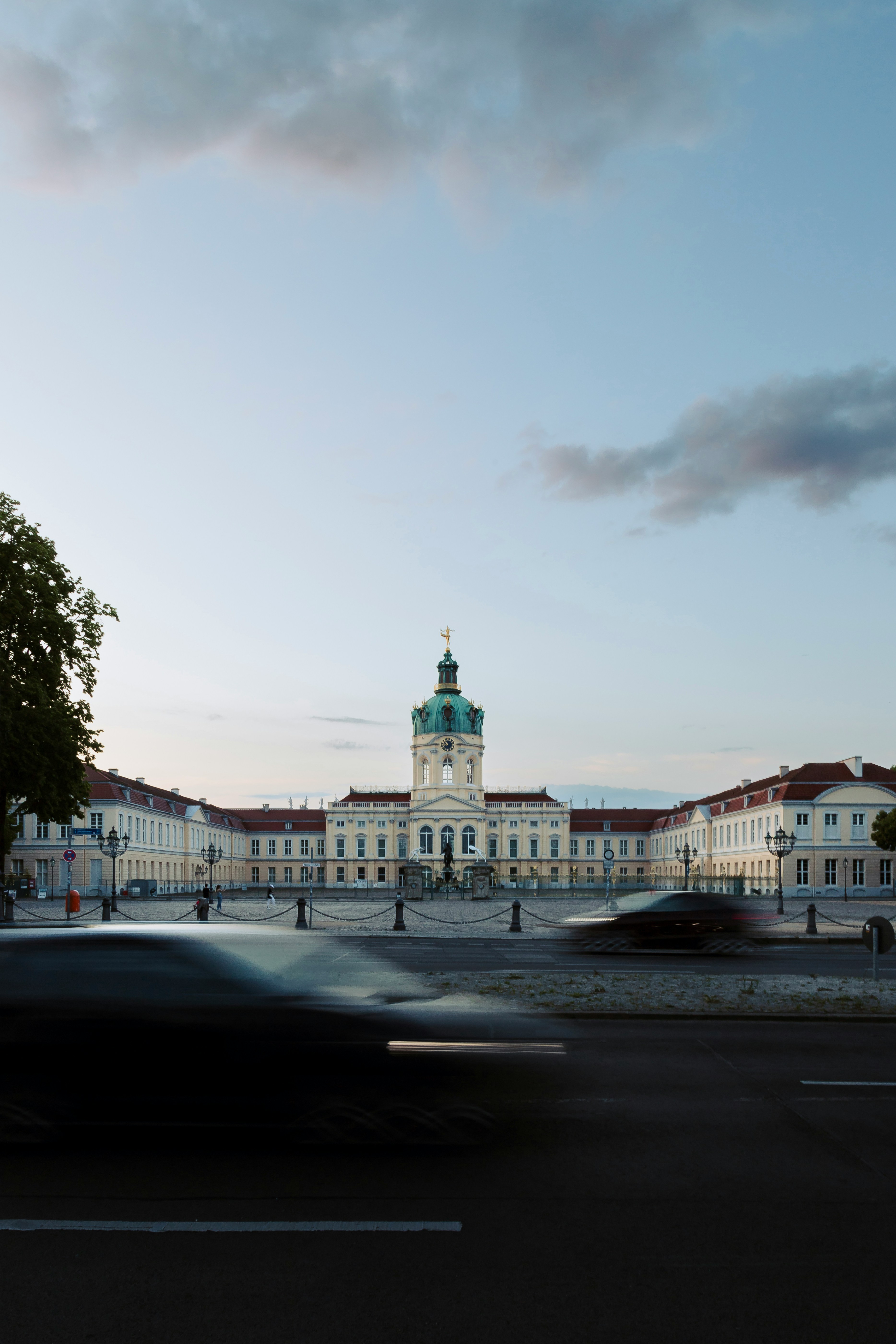 the image shows the charlottenburg palace in berlin, germany, taken from a street level perspective. the palace is a grand baroque building with a central dome and two flanking wings. the sky is overcast, and the lighting is soft and diffused. there are a few cars driving past the palace, creating motion blur.