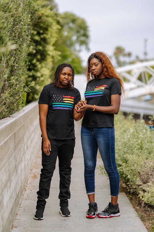 wo young black women, dressed in black tshirts featuring a rainbow flag design and jeans, are walking down a sidewalk, holding hands. they appear to be friends or a couple, exhibiting a sense of togetherness and support. the urban setting is casual, and the overall mood is happy and positive, possibly suggesting a celebration of pride or lgbtq+ identity.