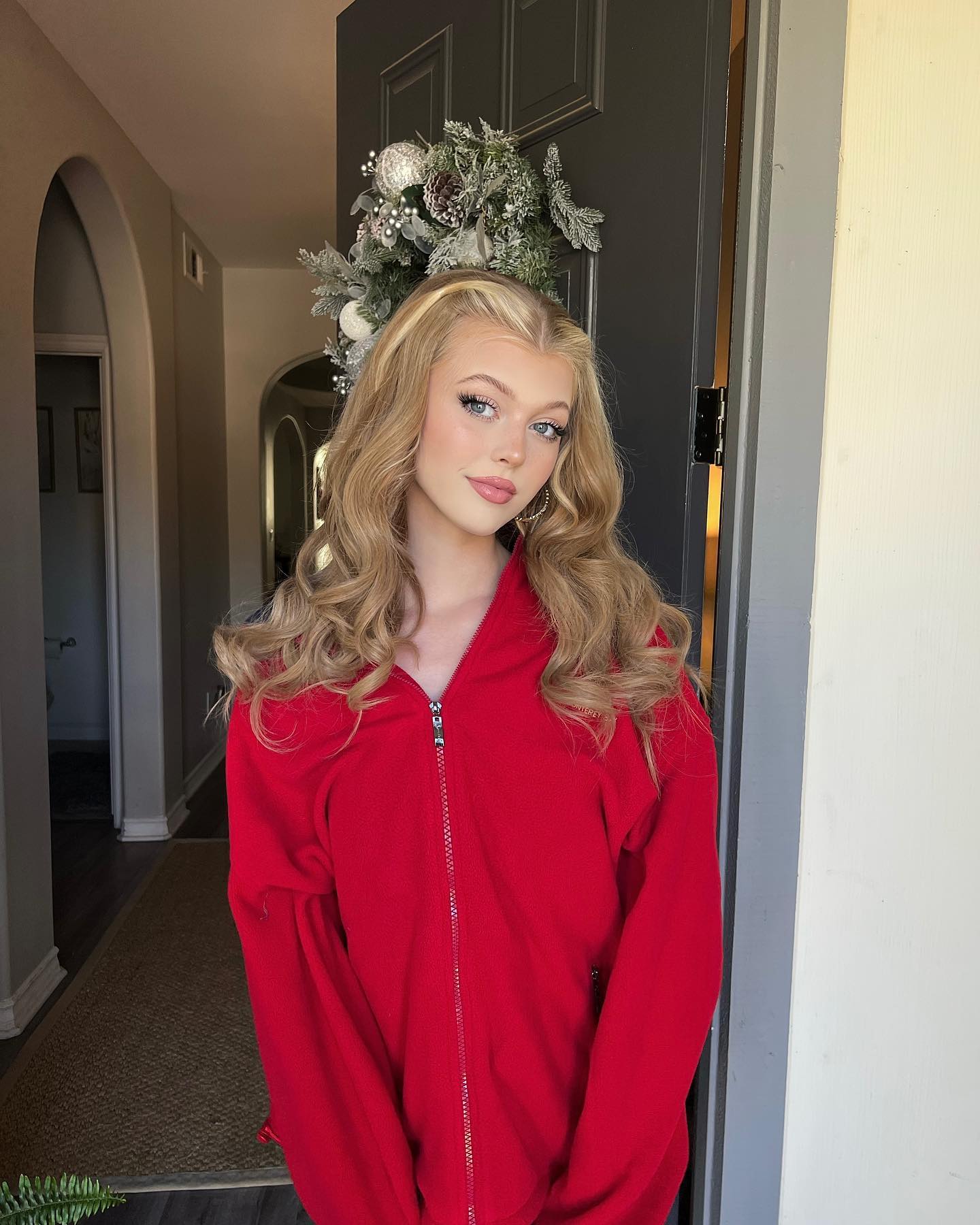a young woman with blonde, curly hair and hoop earrings is standing in a doorway wearing a red zipup jacket. a wreath is hanging on the door behind her. the doorway leads to an interior hallway with an archway.