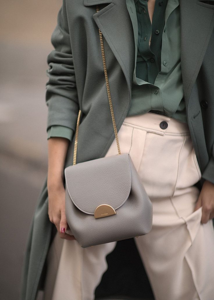 loseup view of a woman wearing a stylish outfit. she is wearing a sage green coat over a sage green silk blouse and a pair of light beige wideleg trousers. a grey leather crossbody bag with a gold chain strap is slung across her body. the overall style is minimalist, sophisticated, and elegant, suggesting a fall or autumn fashion look. the image focuses on the textures and colors of the clothing and accessories.