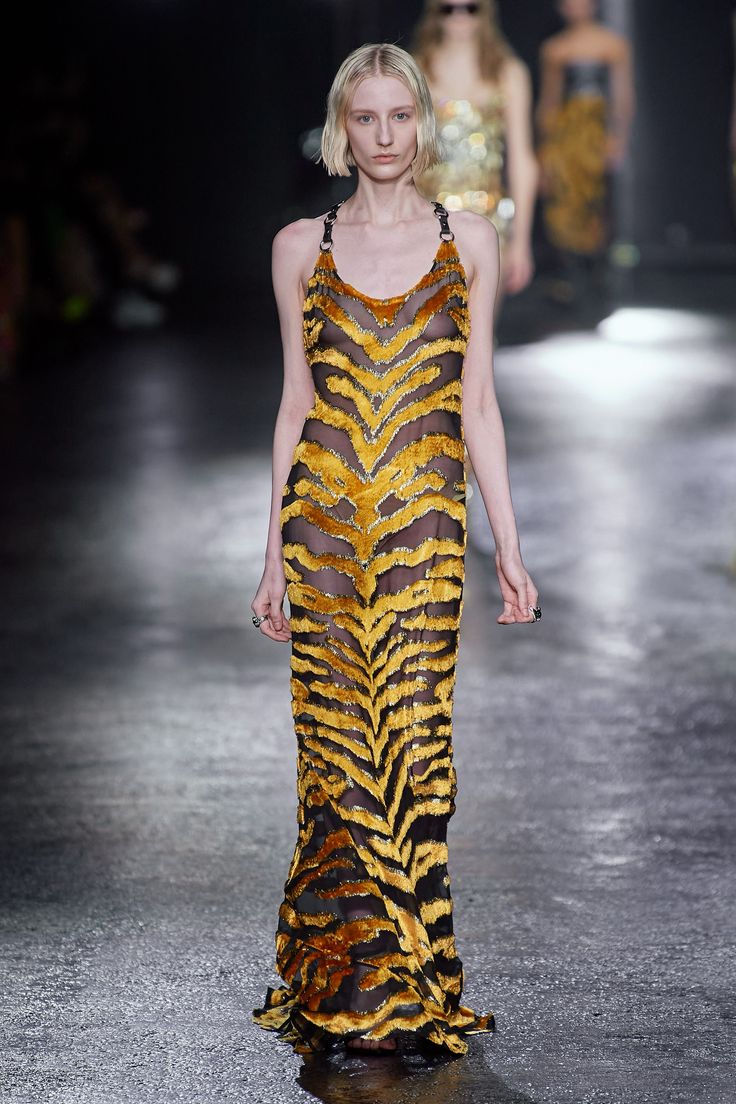 a blonde model walks the runway during a fashion show. she is wearing a long, sleeveless, haltertop dress with a tiger stripe pattern. the dress is sheer in some areas. she is wearing jewelry, including rings. other models and audience members are visible in the background. the runway is dark and reflective.