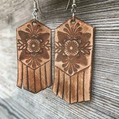 two leather earrings are displayed against a rustic wooden background. the earrings are brown, feature an embossed floral design, and have a fringe at the bottom. the earrings are attached to earring hooks, suggesting they are dangle earrings. they appear to be handcrafted with a rustic aesthetic.