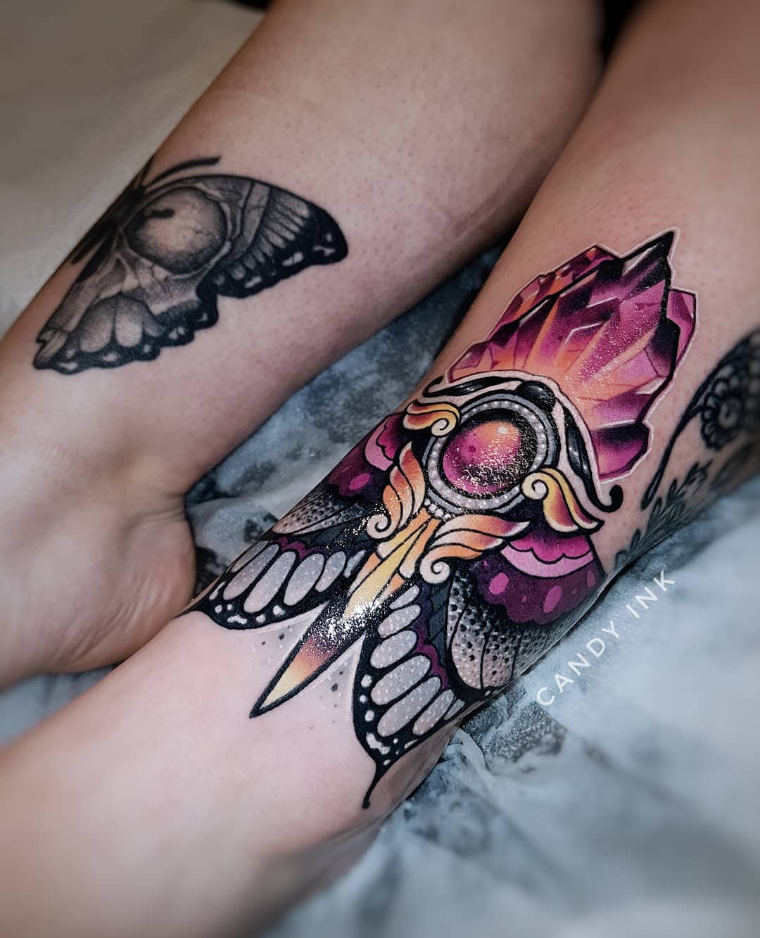 a closeup photograph showcasing two tattoos on an individual's legs. the first tattoo, located on the calf, depicts a stylized moth with a crystal and dagger design incorporated into its body. the moth's wings are spread, revealing intricate patterns and shading.  the second tattoo, situated on the ankle, features a black and grey butterfly with delicate details.
