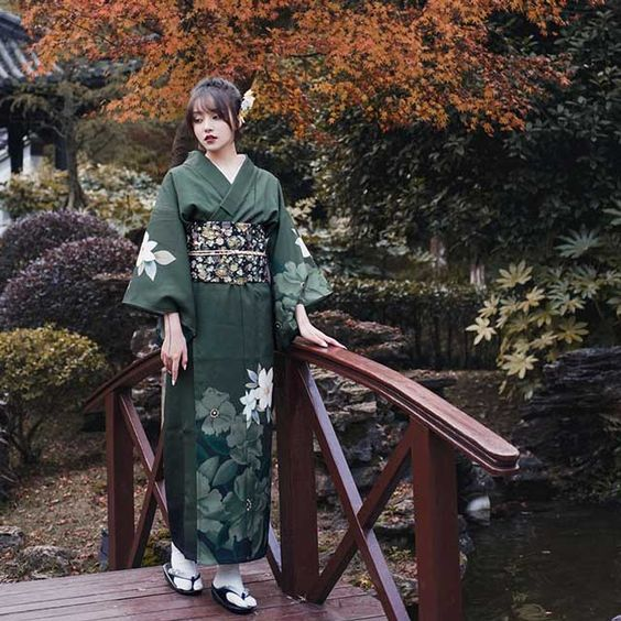a woman in a green kimono with a white floral pattern stands on a wooden bridge in a garden. the garden is lush and green with red and orange leaves on the trees in the background, suggesting it's autumn. the photo is taken from an eyelevel perspective and the lighting is soft and natural.