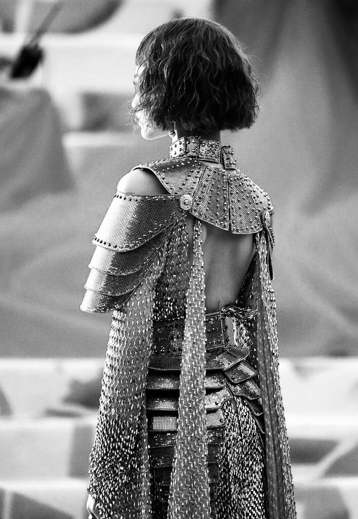 the image is a black and white, closeup shot of a woman from the back. she is wearing an elaborate, silver metallic gown that resembles armor or chainmail. the dress features a capelike back piece and shoulder armor. her hair is a short, wavy bob. the setting appears to be an outdoor event, possibly a red carpet or the met gala, suggested by the blurred background. the overall aesthetic is high fashion, elegant, and striking. the focus is on the intricate detail of the dress and the woman's stylish presentation.