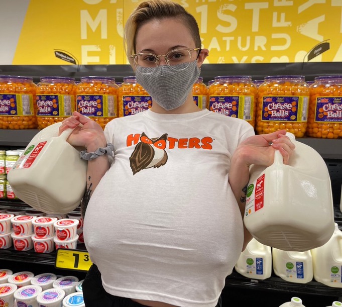 a young woman, wearing a white hooters tshirt and a gray face mask, is seen in a grocery store.  she is holding two large jugs of milk and is standing in front of shelves stocked with cheese balls.  the woman has large breasts and is wearing glasses. the overall image is informal and depicts a casual shopping trip.