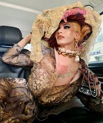 a glamorous drag queen is seated in the backseat of a car. she is wearing an elaborate gold lace outfit with a large matching hat adorned with pink flowers. she has a rose tattoo on her chest and is wearing a substantial amount of jewelry including a large statement necklace.  she is holding a small black handbag.  the overall impression is one of high fashion and theatrical flair.
