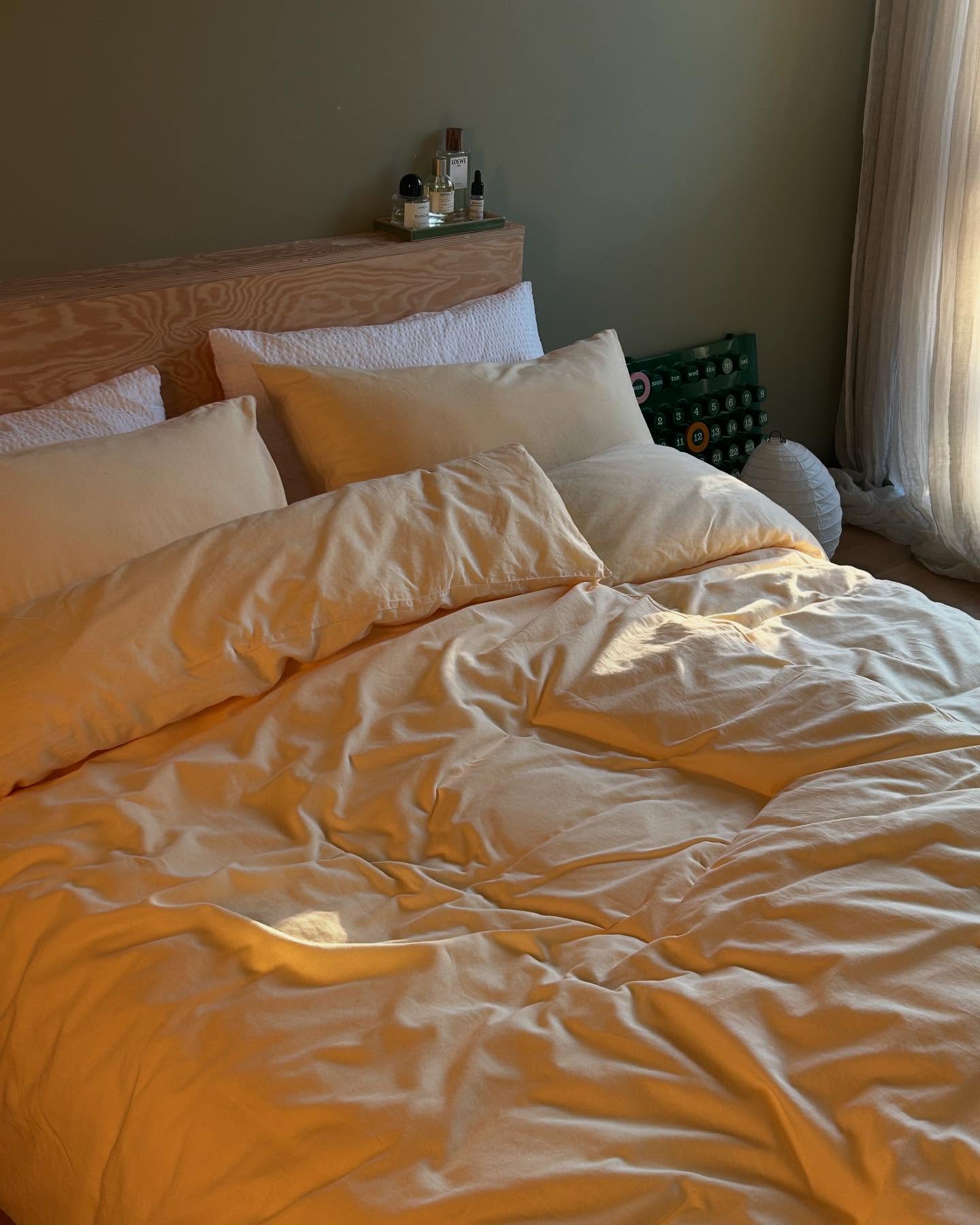 a bed with yellow bedding and pillows is visible in the image. there is a wooden headboard behind the pillows. a curtain hangs on the right side of the image, and there is a lamp on a nightstand behind the pillows. there are also bottles and skincare products on the nightstand. the wall behind the bed is painted in a dark green color.