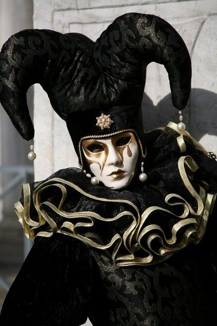 a person wearing a jester costume, hat, and mask during a masquerade ball. the costume is black with gold accents and the mask is white with gold paint. the jester costume is made with a ruffle collar and hat with horns.