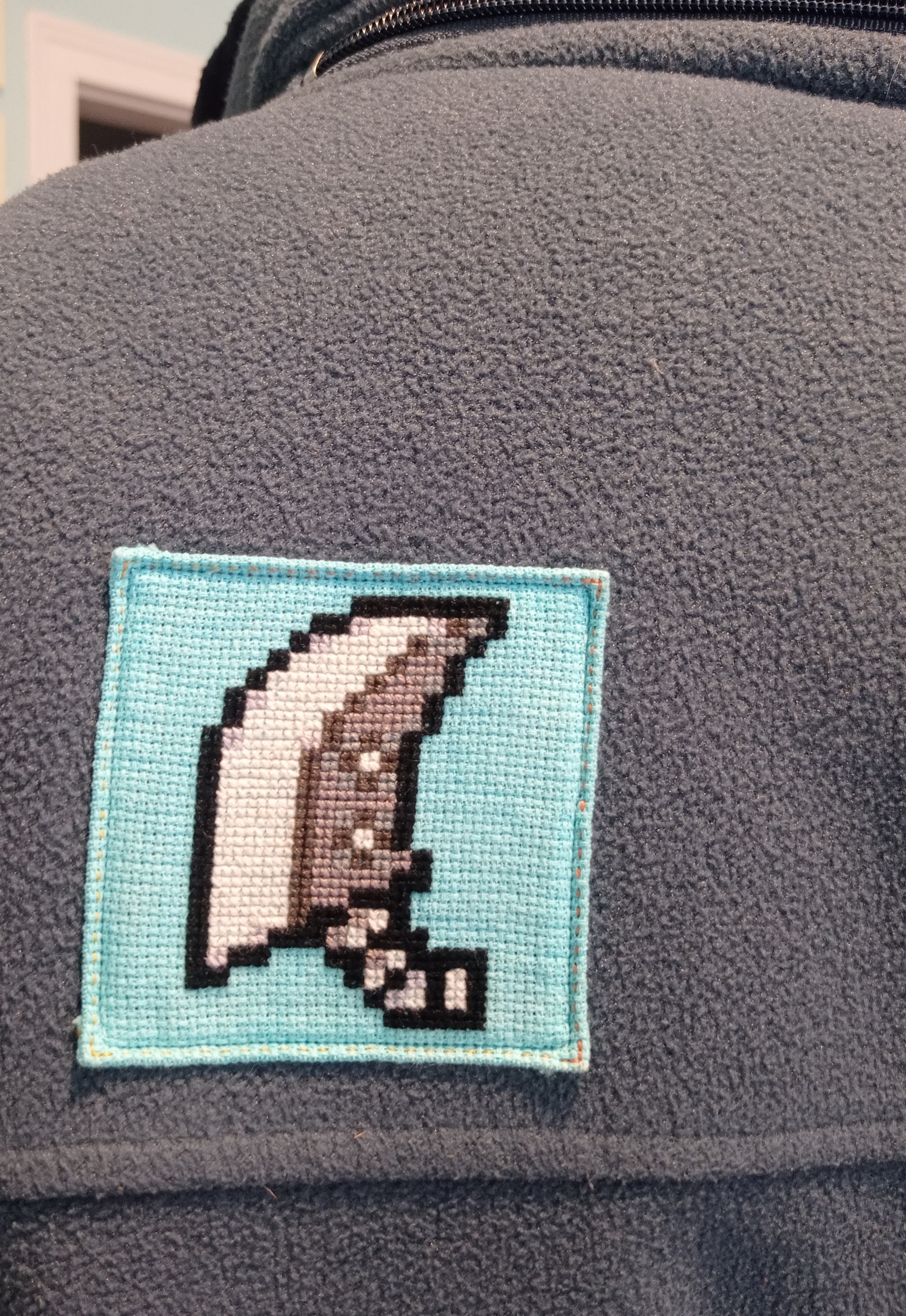 loseup view of a dark grey fleece jacket with a small, square crossstitch patch sewn onto it. the patch features pixel art depicting a crescent moon in shades of grey and white, set against a light blue background. the fleece texture is visible, and the stitching around the patch is neat. the overall style suggests a handmade or custommade garment.