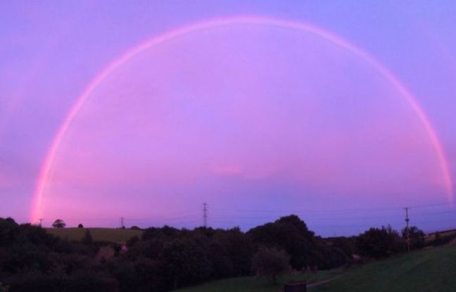 a photograph captures a vibrant pink and purple sunset with a faint rainbow arc in the sky. the horizon is dotted with trees and fields, creating a tranquil and idyllic setting. the soft light and colors evoke a sense of peace and beauty.