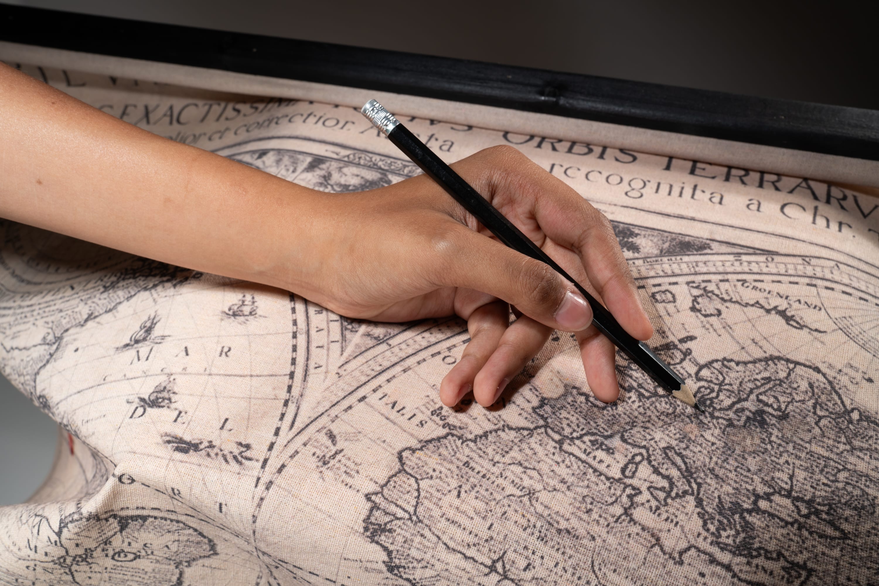 a person's hand is holding a black pencil and pointing at a vintage world map. the map is in black and white with a beige background and features continents, oceans, and geographical details. the image is taken indoors with studio lighting and features a firstperson perspective.
