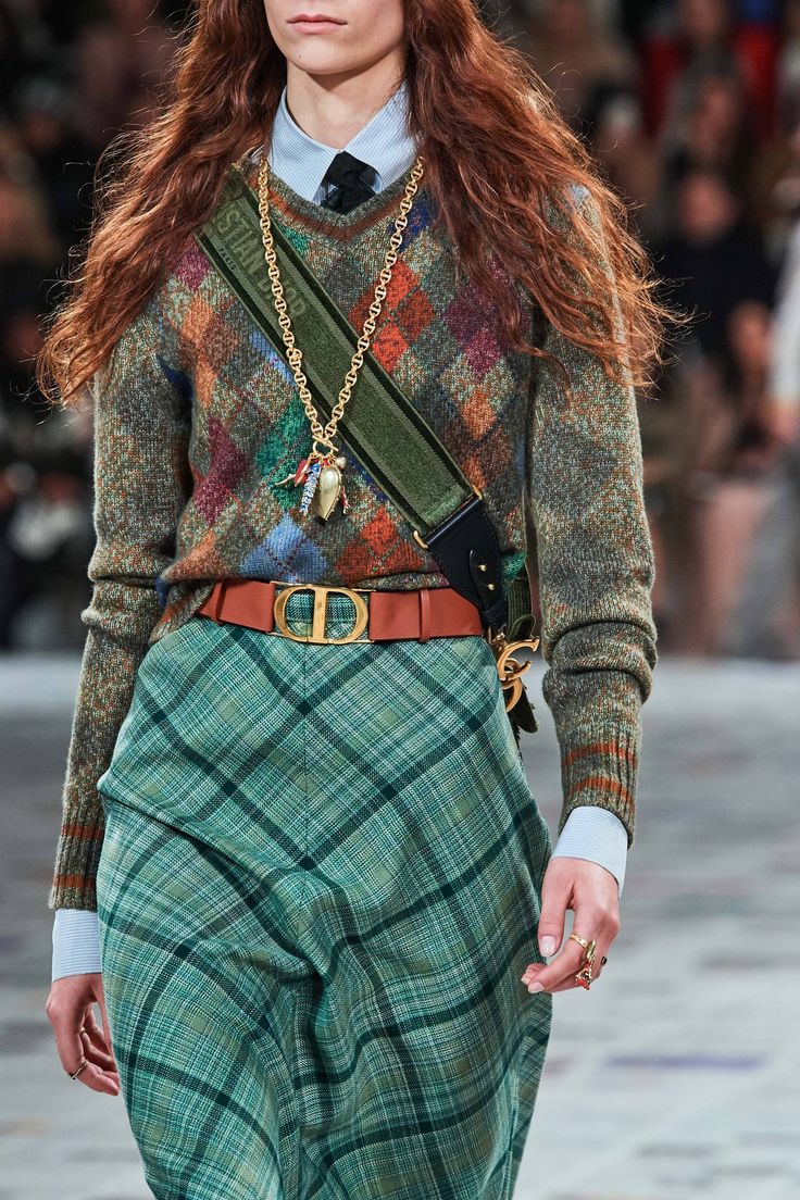 closeup shot of a model on a fashion runway wearing a green and brown plaid skirt, a multicolored argyle sweater, a dior belt, and a gold necklace with a dior strap bag.  the model's hair is long and reddishbrown.  the overall style is autumnal and chic.