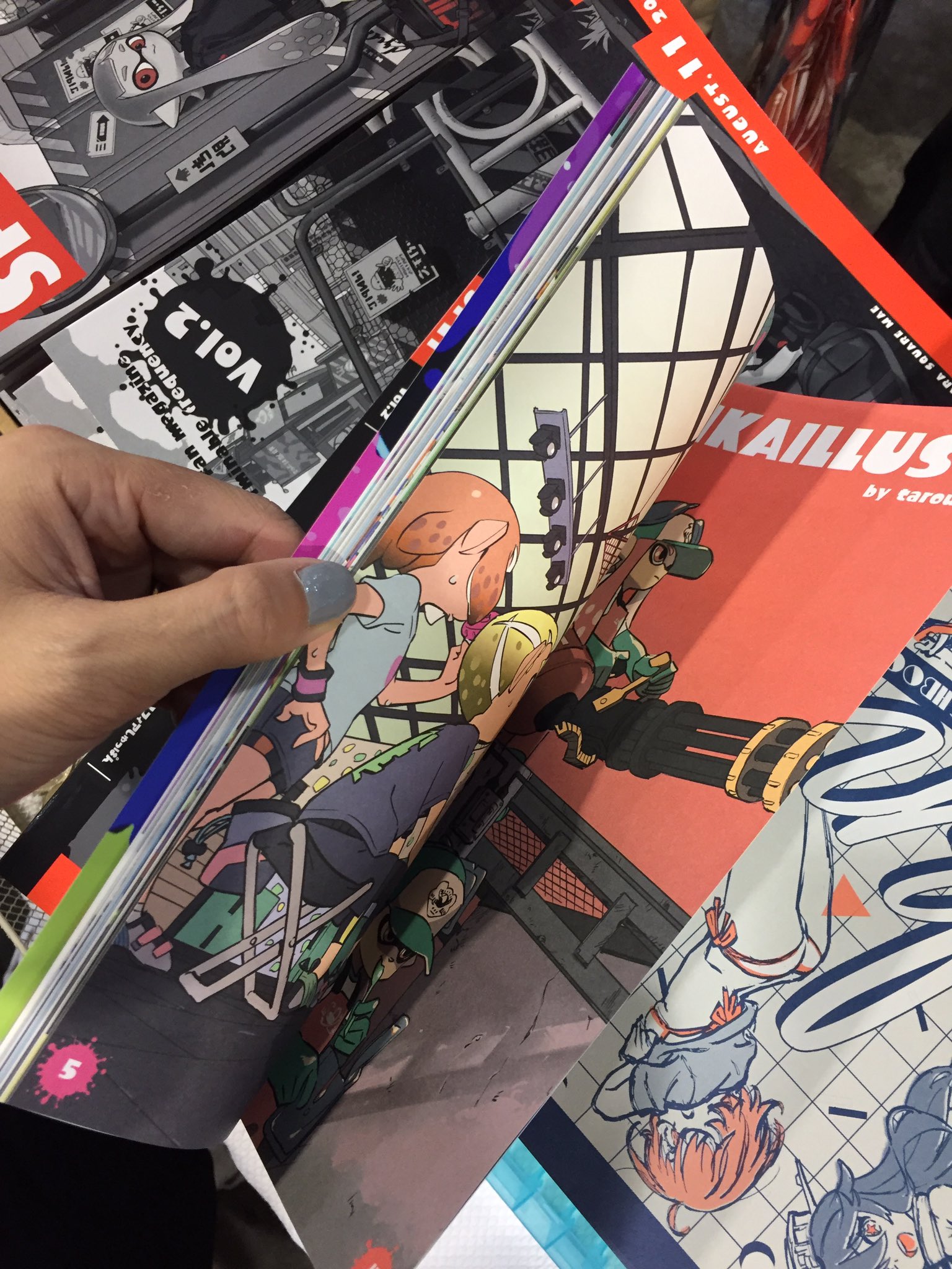 a hand holds up an open comic book, showcasing colorful illustrations of characters that seem to be inspired by the splatoon video game. the book appears to be within a collection of other comics, suggesting a convention or book fair setting.