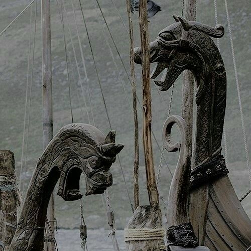 a closeup shot of two intricately carved wooden dragon heads on the bow of a viking ship. the dragon heads face each other with open mouths, revealing detailed teeth and carvings. the ship has visible masts and rigging against a background of cloudy skies.