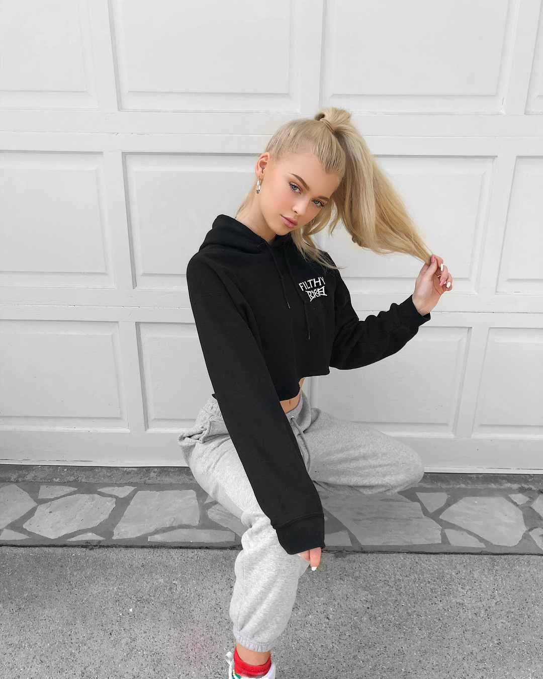 a blonde woman with her hair in a high ponytail is crouched in front of a white garage door. she is wearing a black cropped hoodie and grey sweatpants. she is holding a piece of her hair in her right hand. she is wearing red and white shoes.