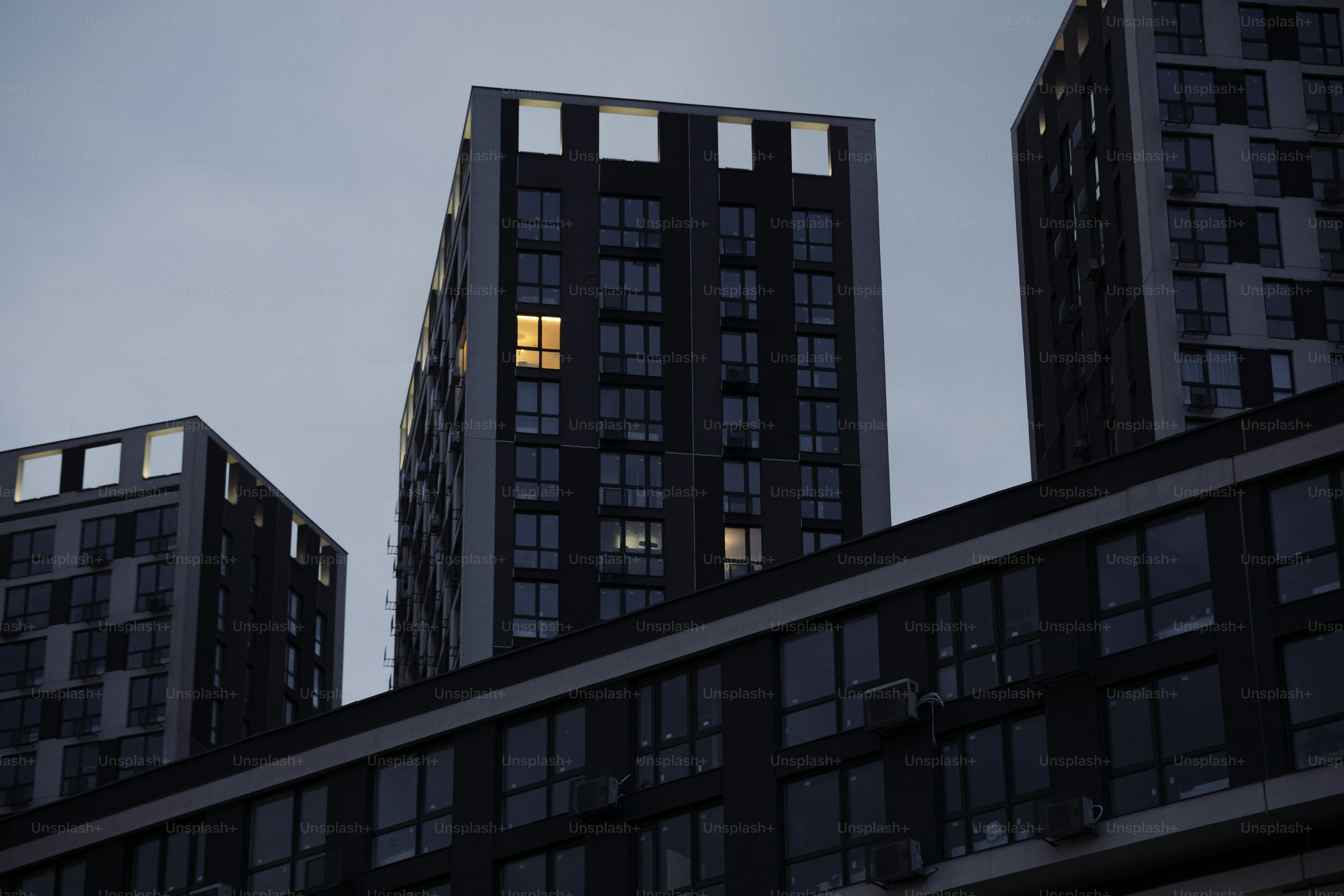 the image shows a lowangle view of several modern apartment buildings. the buildings are dark and imposing against a pale sky, likely at dusk or dawn. the architecture is characterized by clean lines, rectangular windows, and a minimalist design. only a few windows are illuminated, suggesting occupancy. the overall tone is subdued and somewhat stark, emphasizing the scale and uniformity of the urban landscape.