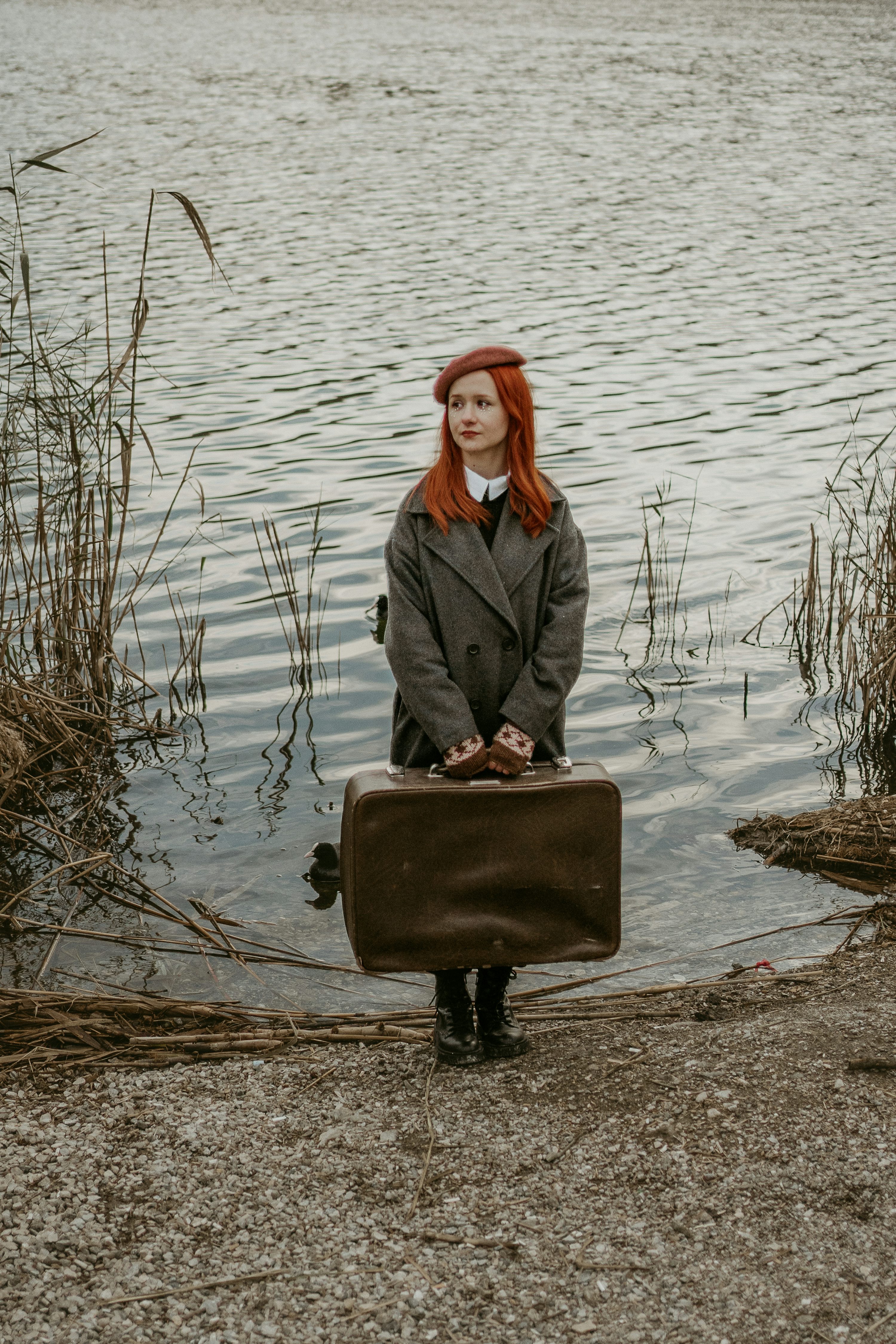 a young redhead woman wearing a beret and a grey coat stands by a lake holding a vintage suitcase. she looks pensive and thoughtful. the atmosphere is autumnal and melancholic. the scene is outdoors, near a lakeshore with reeds and gravel. the overall style is retro and stylish.