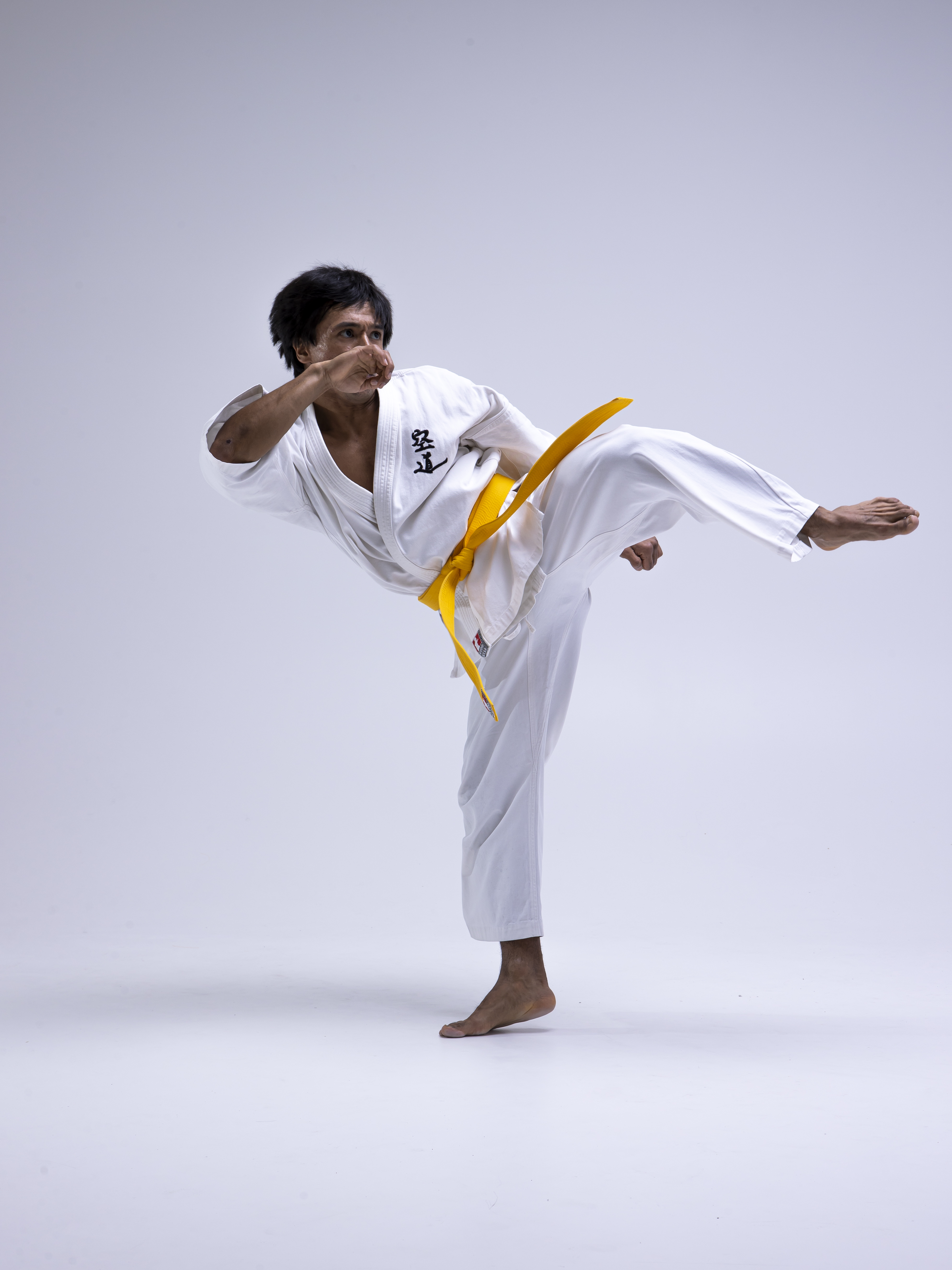 a man in a white karate uniform with a yellow belt is performing a kicking stance on a white background. he is barefoot and appears to be in the middle of a training session. his fist is raised in front of him. the uniform is marked with black symbols.