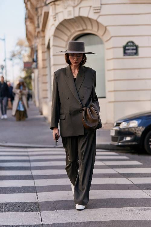 a stylish woman in a doublebreasted grey blazer, matching pants, a fedora hat, white sneakers, and a brown handbag is crossing a pedestrian crossing in a city street. she is walking confidently and looks fashionable. the setting is urban, and the overall style is chic and timeless. the image is welllit and captures the essence of street style photography.