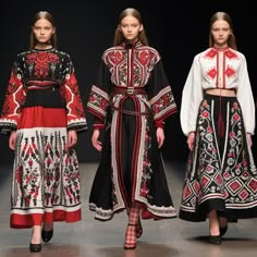 three women in embroidered, stylish clothing stand on a runway. the woman in the middle wears a black and red longsleeved, long dress. the woman on the left is in a red and black skirt and a white, shortsleeved blouse. the woman on the right is wearing a white, shortsleeved blouse and a black, long skirt. all three women have their hair down, and are looking at the camera.