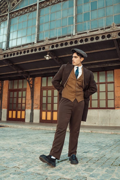 a man stands outdoors wearing a brown suit, vest, and cap. he has his hands behind his back and is looking to the side. behind him is a brick building with large windows and a metal structure. the ground is cobblestone.